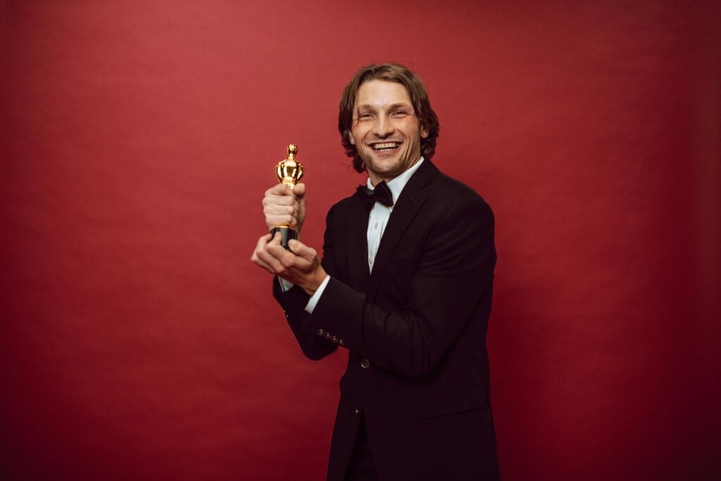 pexels-photo-7005622-7005622 Smiling man in a tuxedo holding a trophy against a red background, celebrating success.